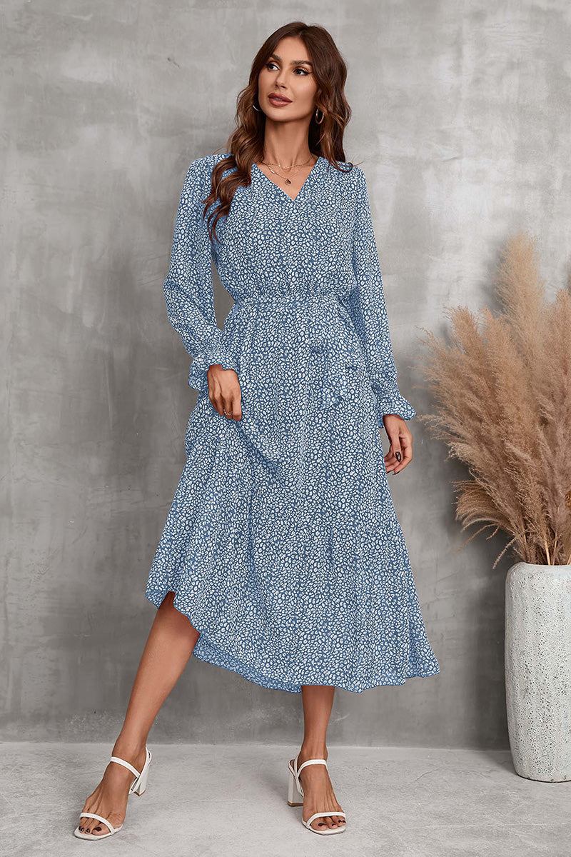 Woman wearing blue Bloom Wrap Midi Dress with long sleeves and floral print standing against gray wall near pampas grass in vase