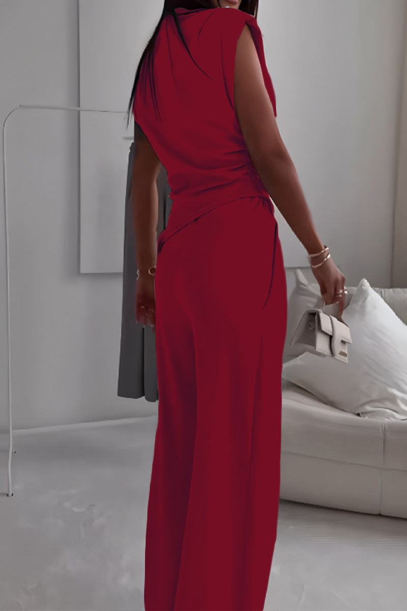 Woman wearing a sleeveless red Manhattan Lines Draped two-piece set with asymmetrical top and wide-leg pants holding a white clutch in a modern room.