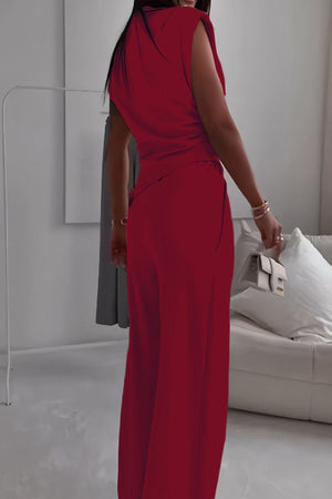 Woman wearing a sleeveless red Manhattan Lines Draped two-piece set with asymmetrical top and wide-leg pants holding a white clutch in a modern room.
