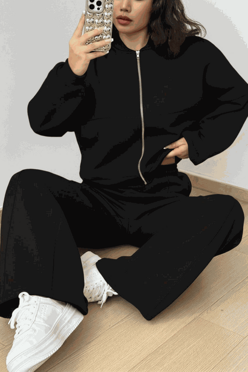 Woman wearing The Voyager Zip Set black zip-front sweatshirt and wide-leg joggers with white sneakers sitting on floor taking a mirror selfie