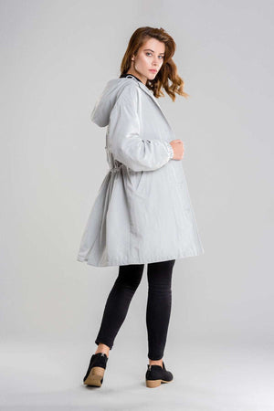 Woman wearing light gray water resistant oversized hooded windbreaker rain jacket with black leggings and shoes standing sideways on gray background