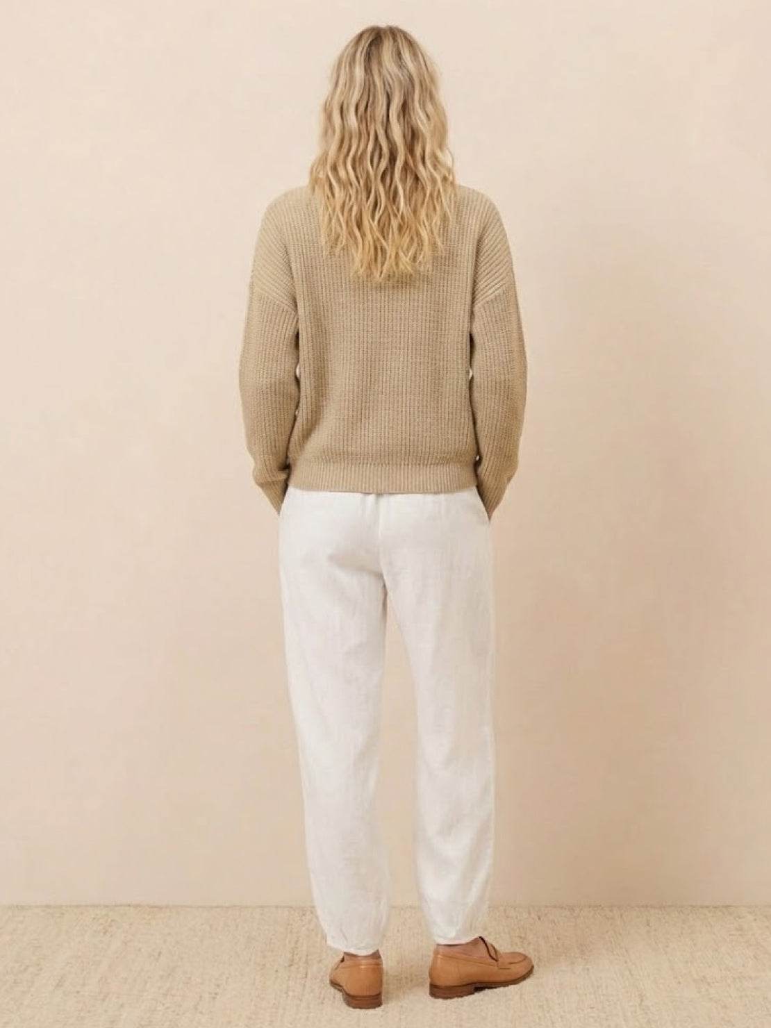 Woman wearing European minimalism relaxed sweater top and white trouser set from the back standing against beige background