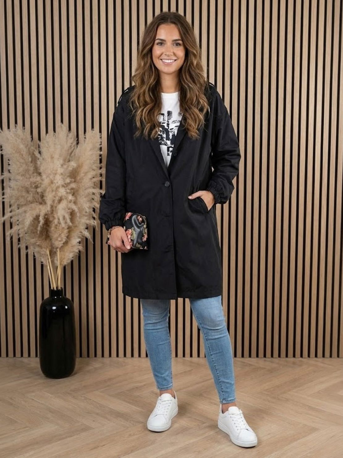 Woman wearing black Rain Couture Windbreaker oversized jacket with jeans and white sneakers standing indoors