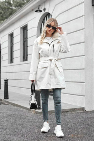 Woman wearing white water-resistant windbreaker trench jacket with belt, sunglasses, jeans, and white sneakers outdoors