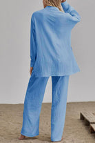 Woman wearing light blue linen blend two-piece set with loose long-sleeve top and wide-leg pants, standing barefoot on sandy floor.