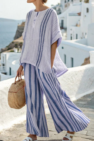 Woman wearing coastal comfort linen two-piece set with blue and white vertical stripes, loose fitting top and wide-leg pants, holding woven handbag outdoors