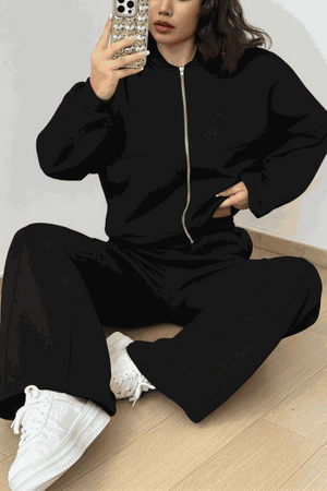 Woman wearing The Voyager Zip Set black zip-front sweatshirt and wide-leg joggers with white sneakers sitting on floor taking a mirror selfie