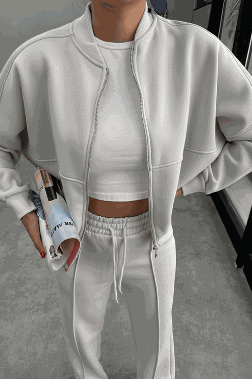 Woman wearing The Voyager Zip Set white zip-front sweatshirt and wide-leg joggers holding a magazine in a modern indoor setting