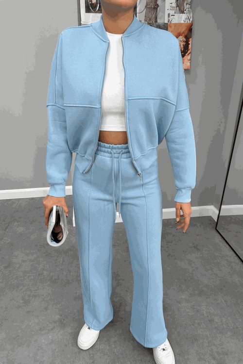 Woman wearing The Voyager Zip Set in light blue, featuring a zip-front sweatshirt and wide-leg joggers with pockets, styled with white sneakers and a white cropped top.