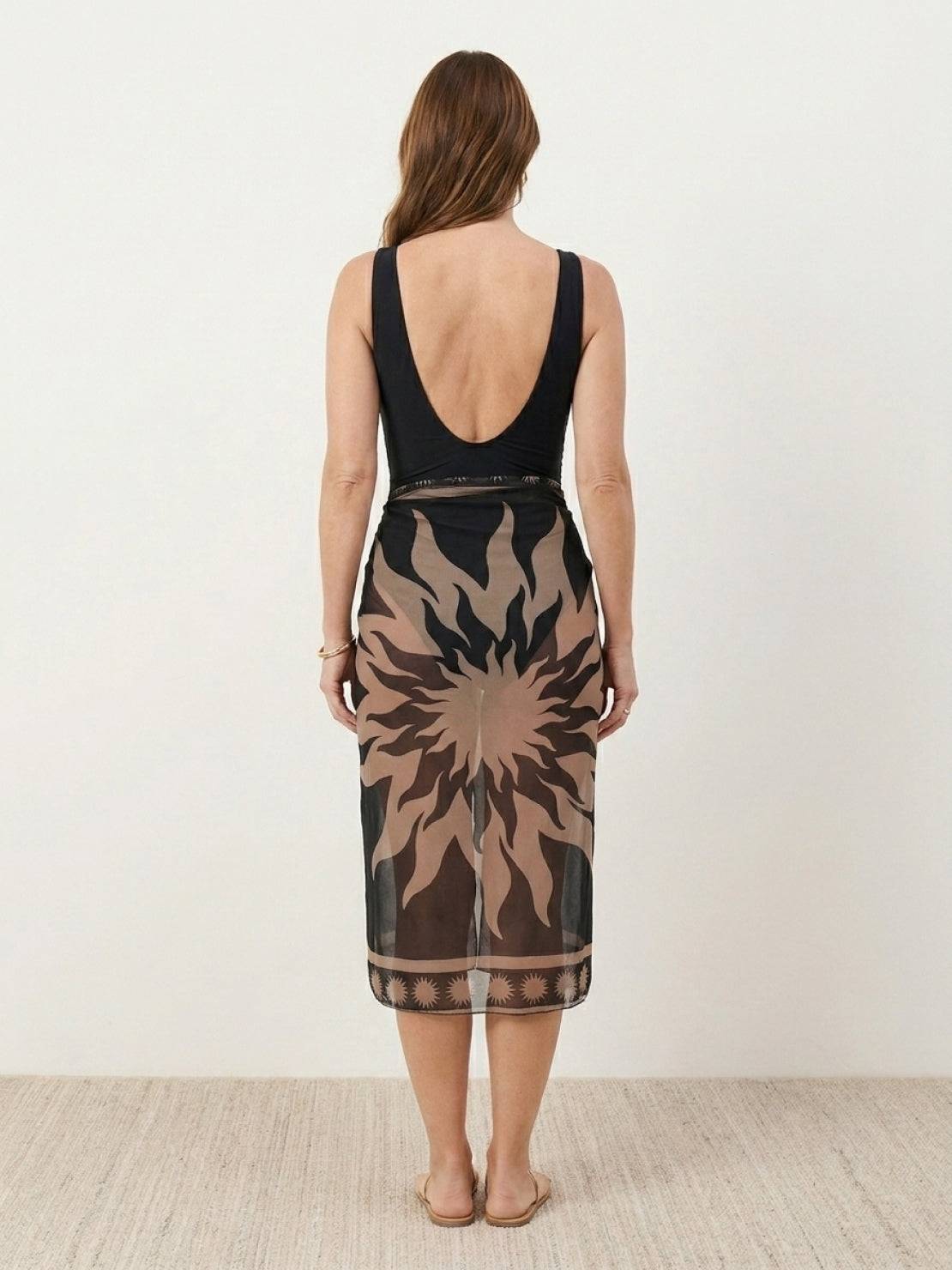 Woman wearing a deep-V swimsuit with ruched bust and waist, paired with a sheer wrap sarong tied at the hip.